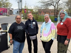 four women standing outdoors in front of tables with garden supplies in an urban neighborhood